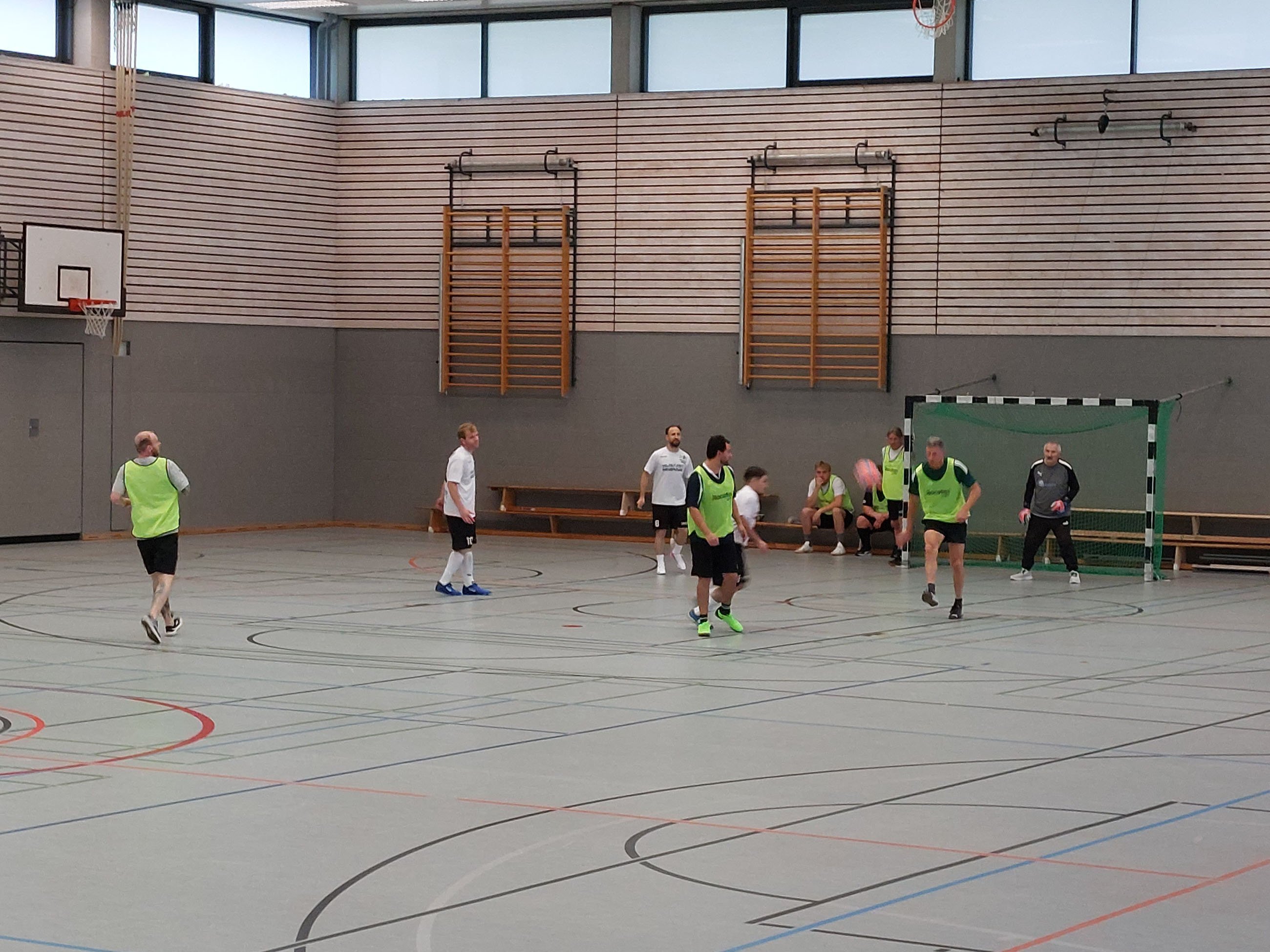 Spielsituation beim 1. Moin Cup in einer Sporthalle in Oldenburg. Mehrere Fußballspieler in weißen Trikots und grünen Leibchen sind auf dem Spielfeld zu sehen. Im Hintergrund steht ein Torwart vor einem Hallentor, während weitere Spieler am Spielfeldrand sitzen.