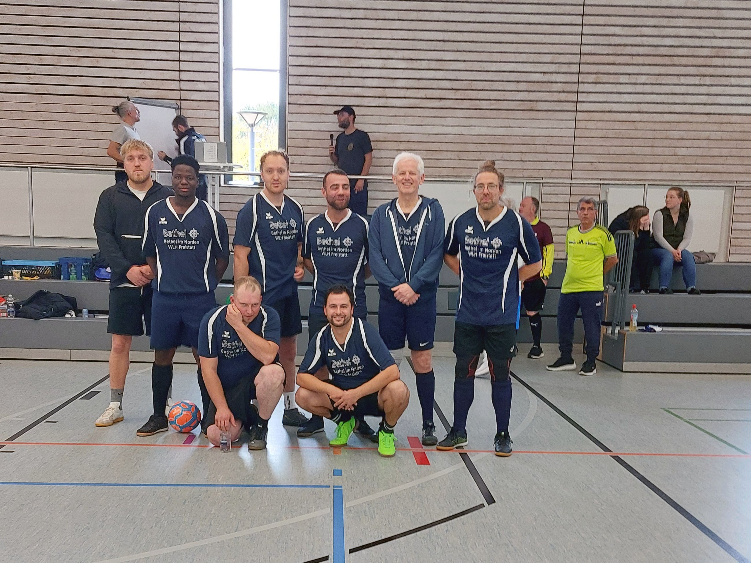 Gruppenfoto der Fußballmannschaft von Bethel Freistatt in einer Sporthalle. Acht Männer posieren in zwei Reihen, sechs davon tragen dunkelblaue Trikots mit der Aufschrift „Bethel im Norden“. Zwei Spieler knien vorne neben einem Fußball, im Hintergrund sind weitere Personen zu sehen.