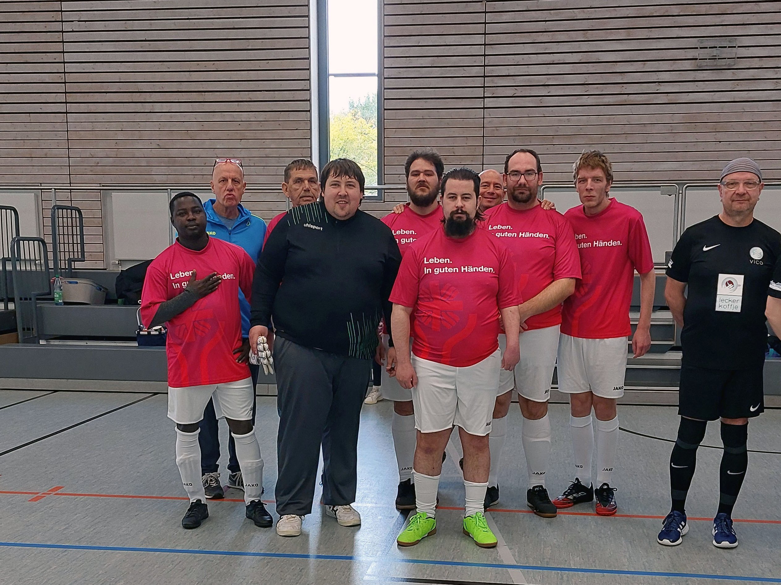 Gruppenfoto der Fußballmannschaft der Caritas Salzgitter in einer Sporthalle. Die Spieler stehen nebeneinander, mehrere tragen rote Trikots mit der Aufschrift „Leben. In guten Händen.“, andere schwarze Sportkleidung.