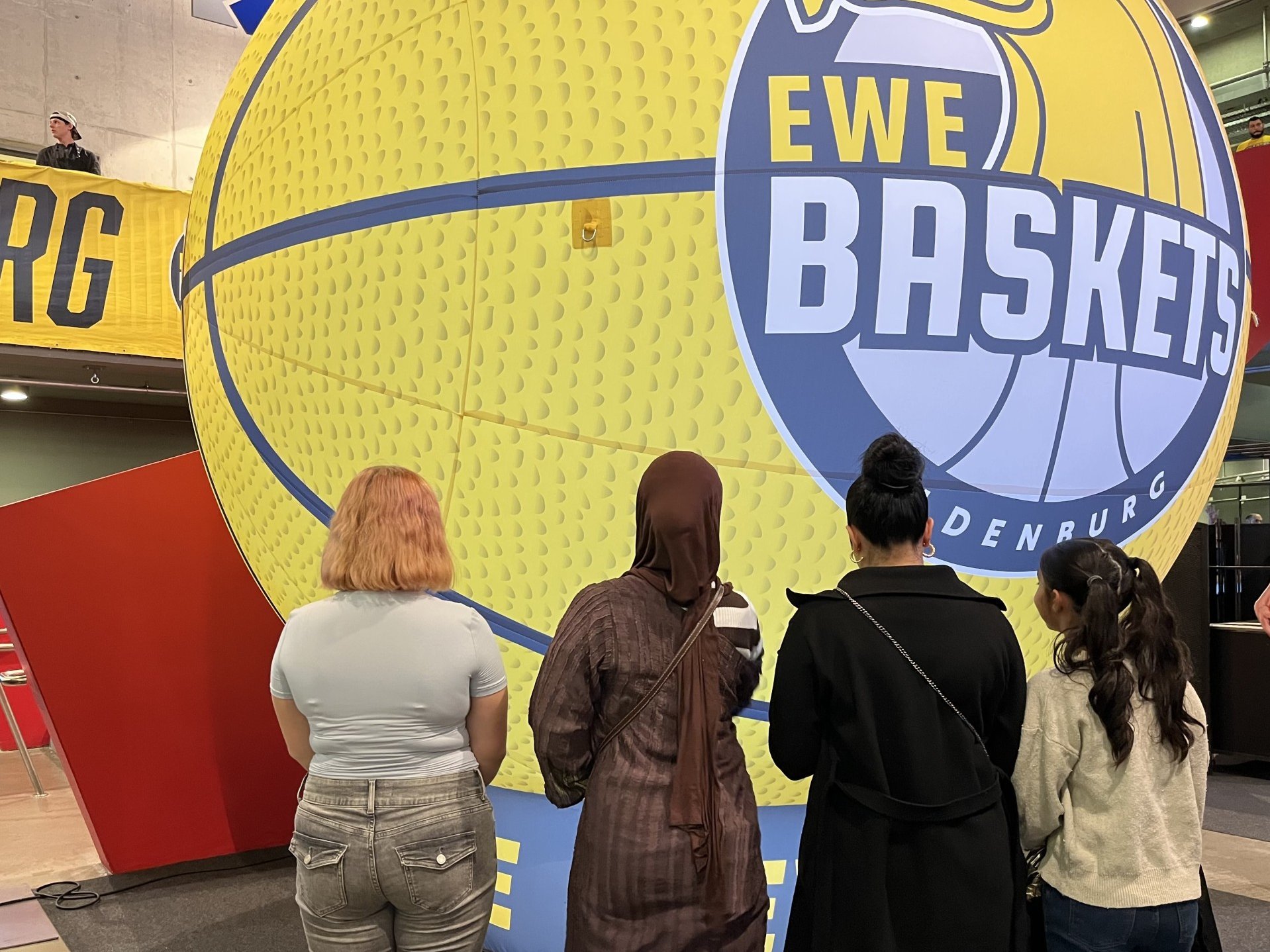 Vier Personen stehen mit dem Rücken zur Kamera vor einem überdimensionalen gelb-blauen Basketball mit dem Logo der EWE Baskets Oldenburg.