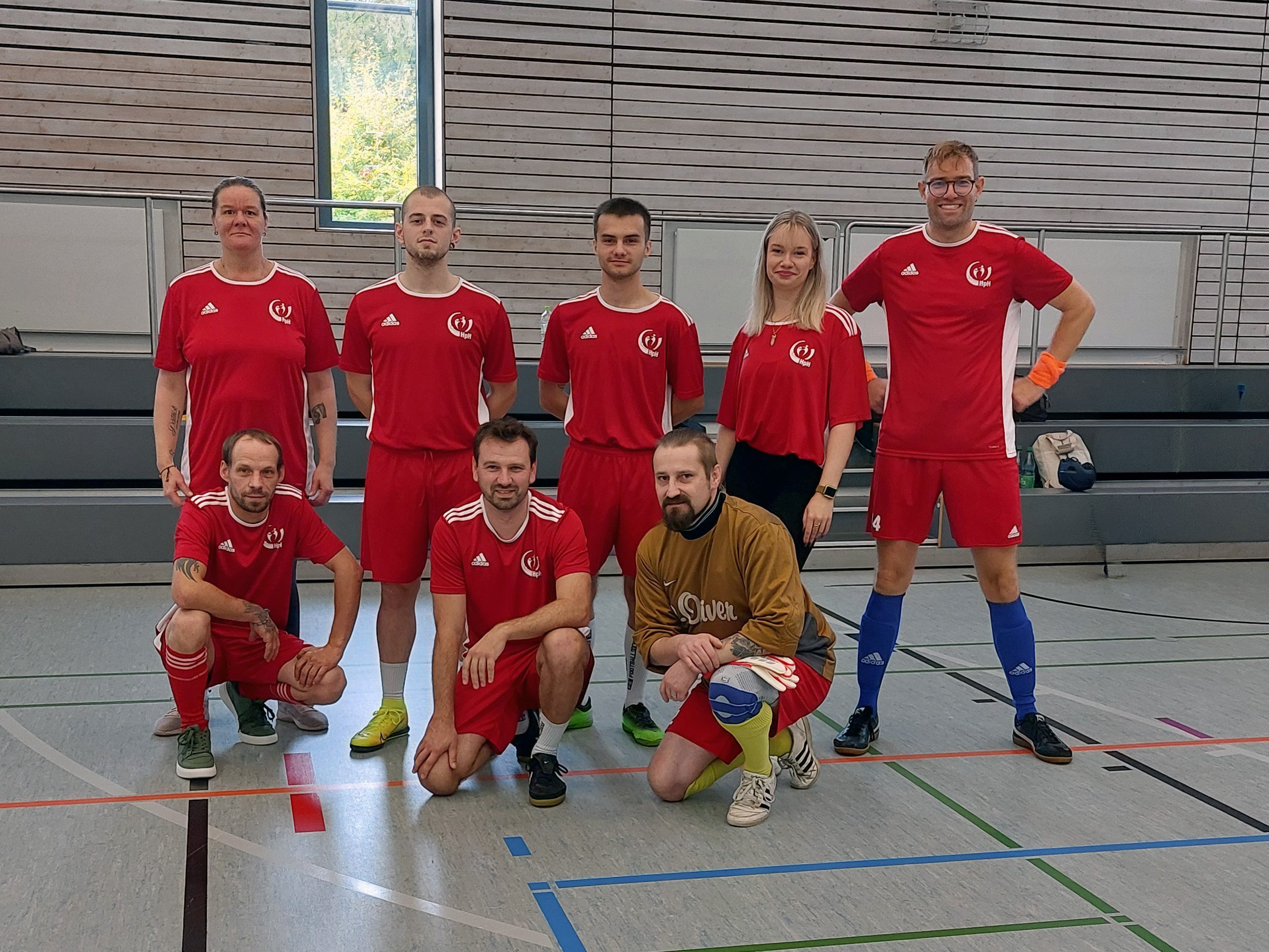 Gruppenfoto der Fußballmannschaft der Heilpädagogischen Hilfe Bersenbrück in einer Sporthalle. Acht Personen posieren in zwei Reihen, fünf stehen hinten und drei knien vorne. Die meisten tragen rote Fußballtrikots. Vorne rechts kniet ein Torwart mit Handschuhen und einem braunen Oberteil.