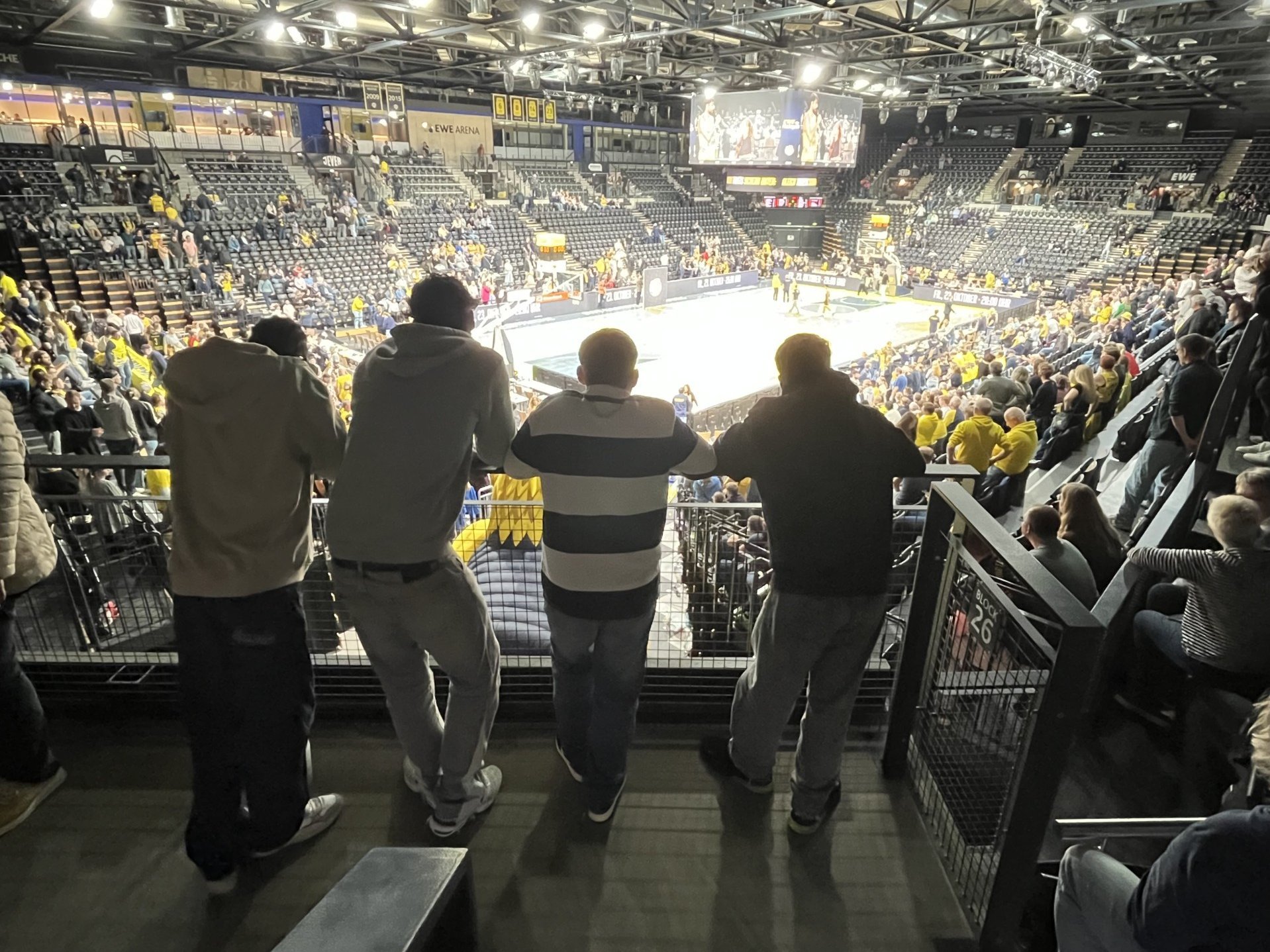 Vier Zuschauer stehen mit dem Rücken zur Kamera an einer Tribünenbrüstung und blicken auf das Basketballfeld in der EWE Arena.