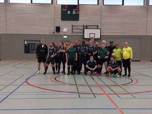 Gruppenfoto der beiden Finalmannschaften Bethel Freistatt und Karl-Jaspers-Klinik (KJK) beim 1. Moin Cup in Oldenburg. Die Spieler stehen und knien gemeinsam in der Mitte einer Sporthalle. Sie tragen überwiegend dunkelblaue und dunkelgrüne Fußballtrikots. Rechts stehen zwei Personen in gelben Schiedsrichtertrikots.