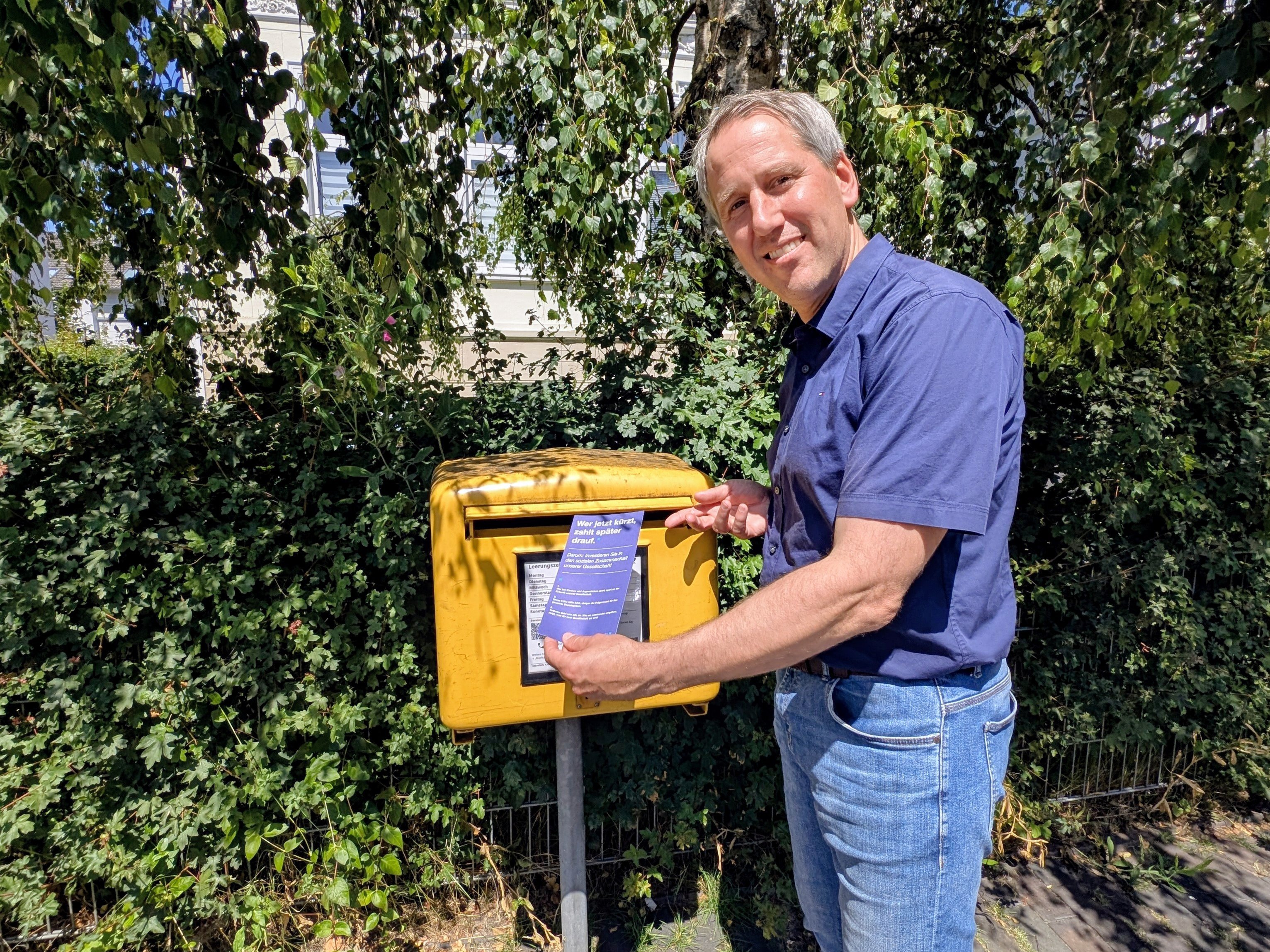 Mario Behrends, Vorstand der Diakonie im Oldenburger Land, wirft eine blaue Aktionspostkarte der Diakonie Deutschland in einen gelben Briefkasten ein. Die Aufnahme entstand an einem sonnigen Tag vor einem großen Baum und verdeutlicht die Beteiligung an der bundesweiten Protestaktion gegen geplante Kürzungen in der Kinder-, Jugend- und Eingliederungshilfe.