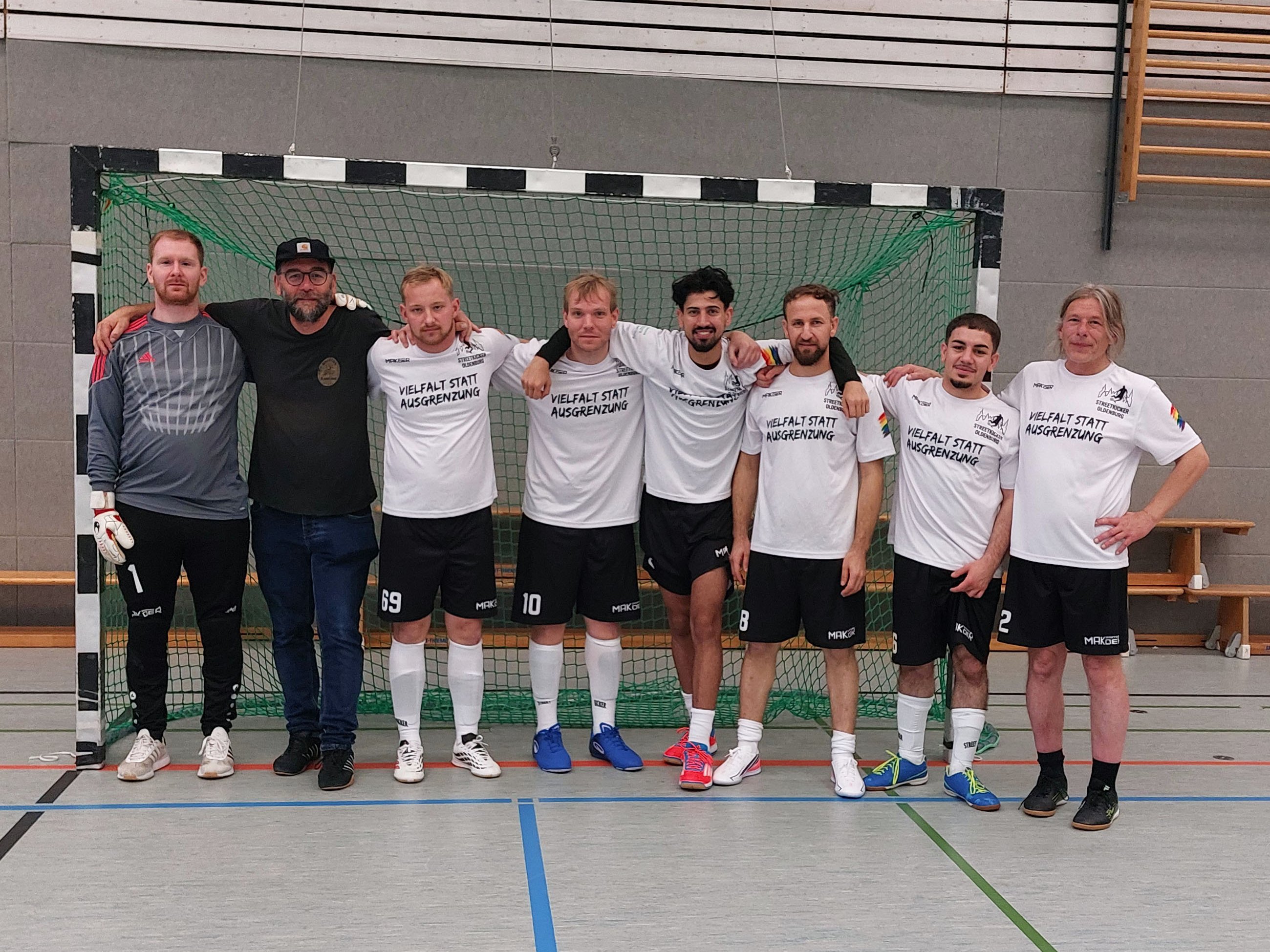 Gruppenfoto der Streetkicker Oldenburg vor einem Fußballtor in einer Sporthalle. Acht Männer stehen nebeneinander, die meisten tragen weiße Trikots mit der Aufschrift „Vielfalt statt Ausgrenzung“ und schwarze Sporthosen. Mehrere Spieler haben die Arme um die Schultern ihrer Mitspieler gelegt.