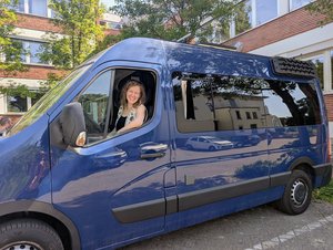 Gesche sitzt lächelnd am Steuer ihres blauen Bullis und schaut aus dem geöffneten Fahrerfenster.