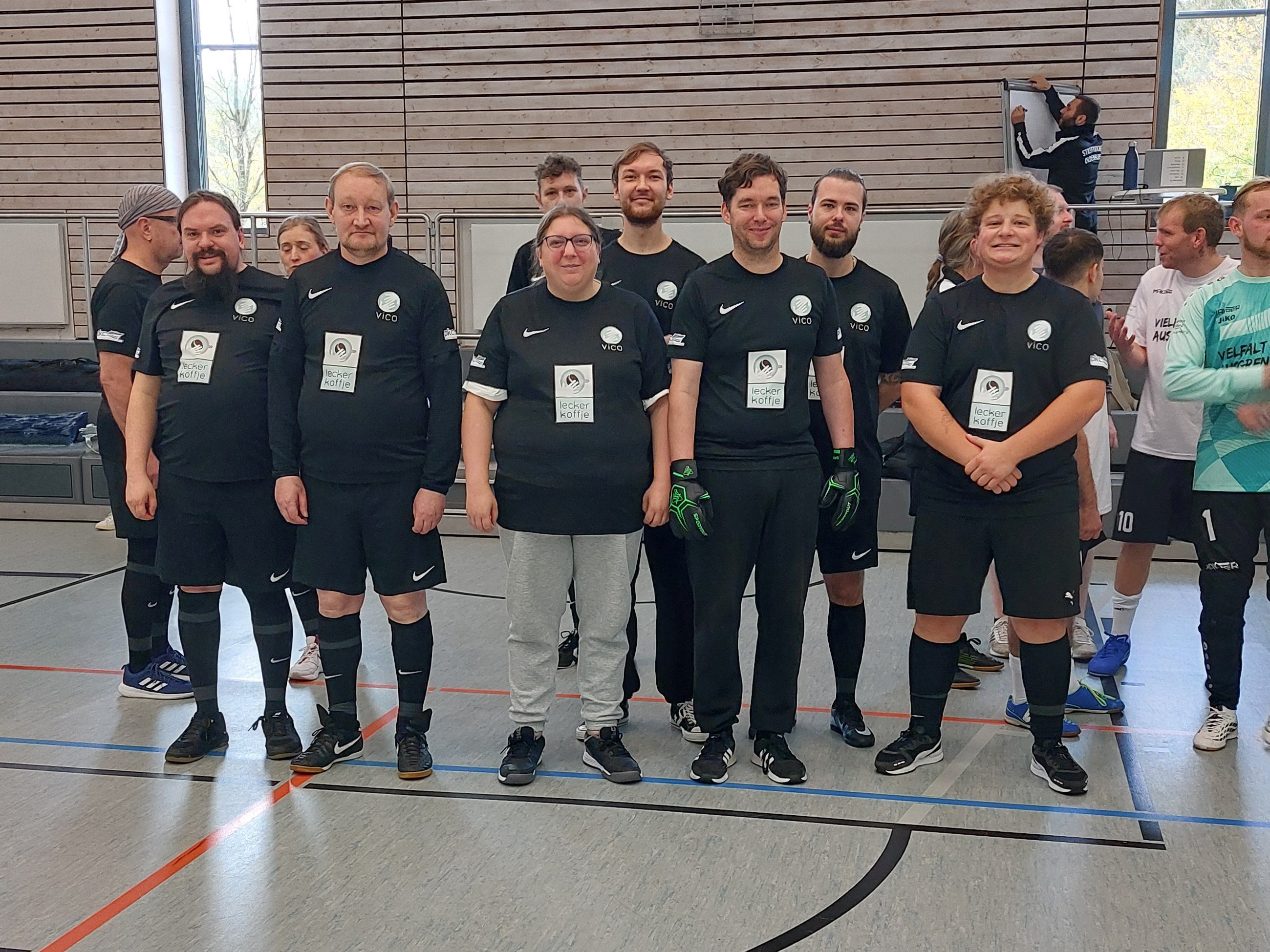Gruppenfoto der Mannschaft der vico GmbH in einer Sporthalle. Acht Personen stehen nebeneinander und tragen überwiegend schwarze Fußballtrikots mit dem vico-Logo und dem Aufdruck „lecker kaffee“. Im Hintergrund sind weitere Teilnehmende des Turniers zu sehen.