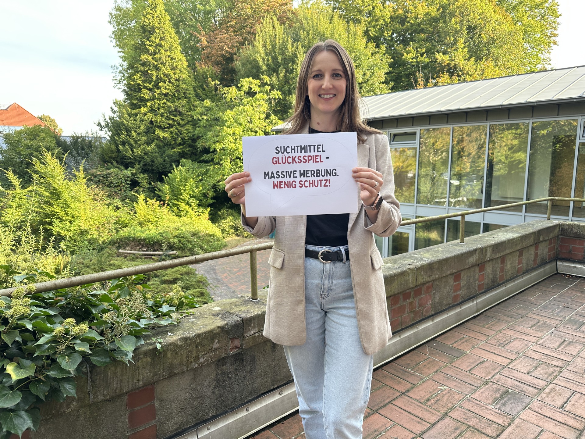Polina Lehmann, Fachkraft für Glücksspielprävention und -beratung bei der Diakonie, hält ein Schild mit der Aufschrift ‚Suchtmittel Glücksspiel – Massive Werbung. Wenig Schutz!‘ in die Kamera.