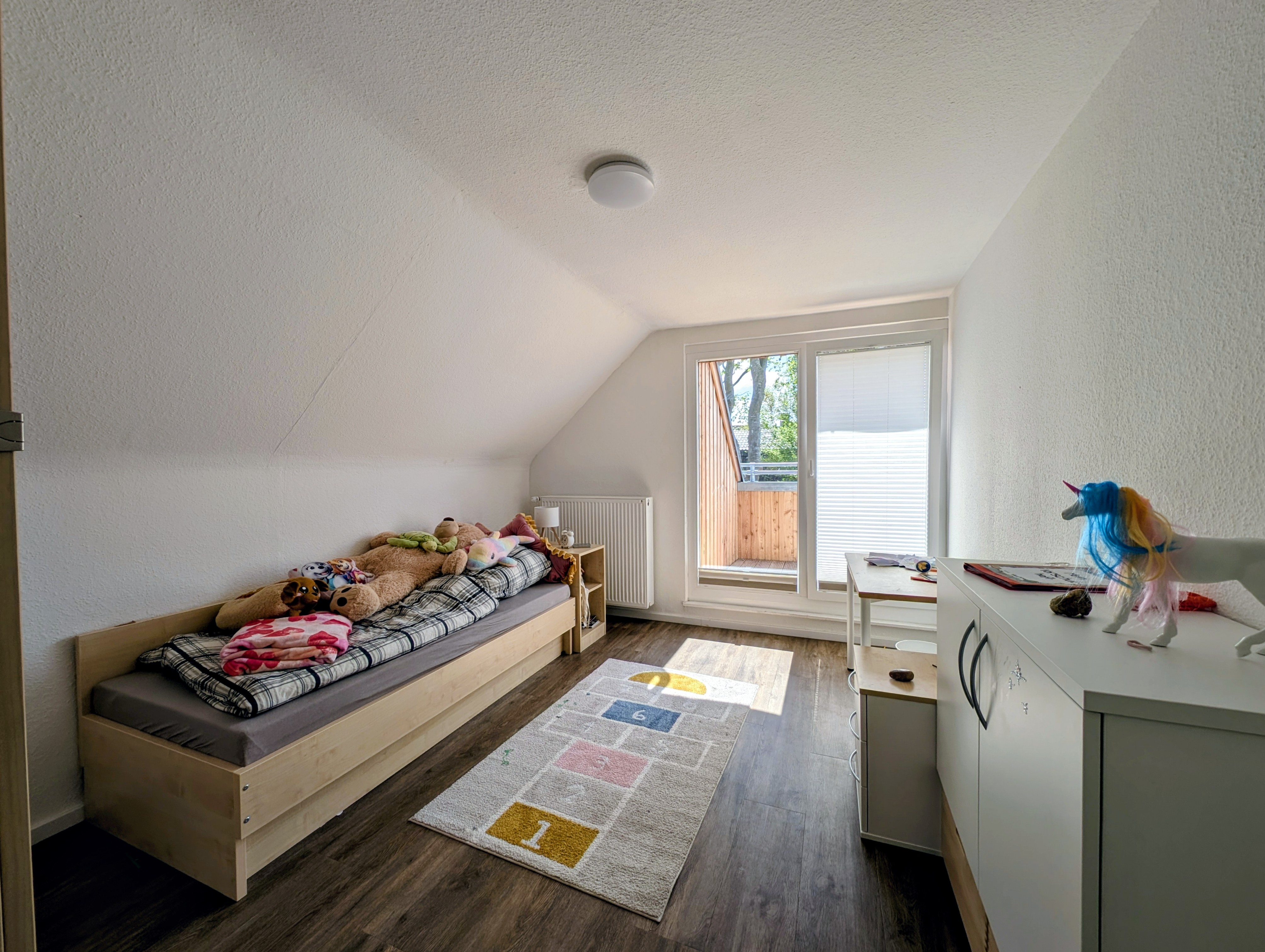 Helles Kinderzimmer mit Einzelbett, mehreren Kuscheltieren, einem kleinen Schreibtisch und einer Balkontür.